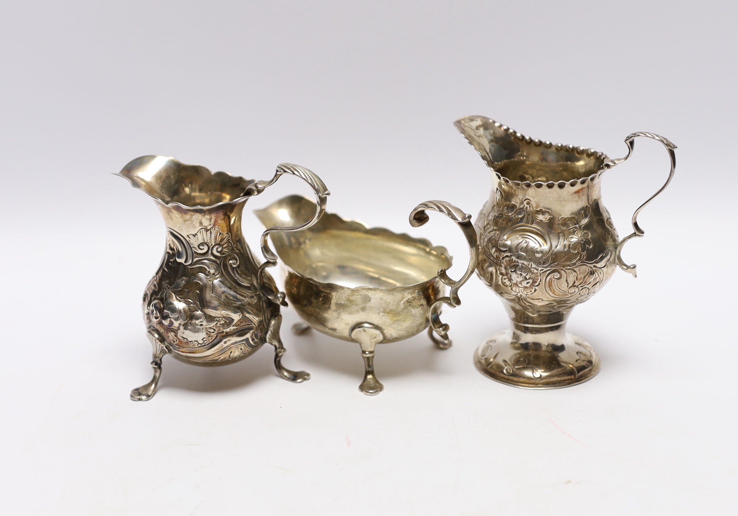 A late George II silver sauceboat with flying scroll handled, London, 1759 and two Georgian silver cream jugs, with later embossed decoration (a.f.)